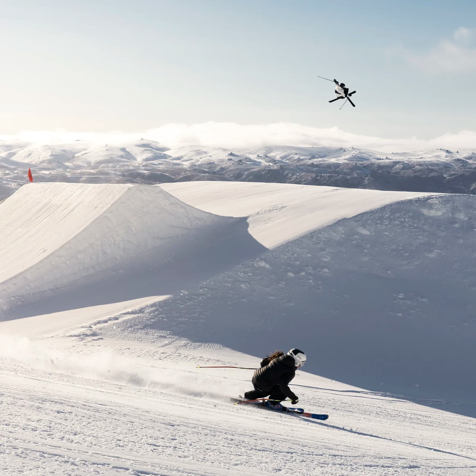 Skier racing down a groomed slope at Cardrona Alpine Resort with a stunning backdrop of snow-covered mountains, while another skier performs an aerial trick in the distance above a large snow jump.